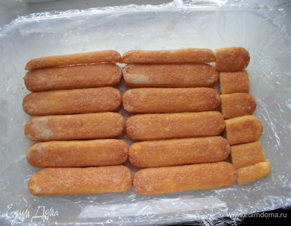 Дно и стенки пластиковой формы (у меня 27 х 20 см) выстелить пищевой пленкой. Важно сделать небольшие концы, которые должны свисать с формы, чтобы потом легче было вынуть торт. Слегка подогреть оставшиеся 90 мл молока, добавить 1 ст. л. сахарной пудры. Выложить печенье в форму. Обильно пропитать его молоком с помощью кисточки. 