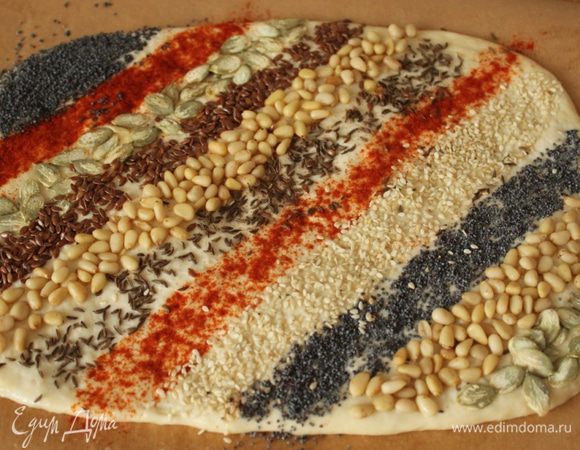 Слегка смазать пласт водой, посыпать тмином, паприкой, измельченными орехами, кунжутом, маком, семечками. Слегка прижать присыпку, пройдясь скалкой.