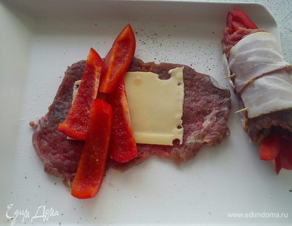 Пластинку сыра и нарезанный полосками перец выложить на мясо. Свернуть рулетик так, чтобы перец выглядывал с обеих сторон. Обернуть рулетик беконом и скрепить зубочисткой