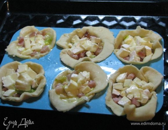 Спустя пол-часа достать из холодильника наши подмороженные тарталетки. Выложить в них начинку: пережаренный бекон с луком пореем, сыр бри, порезанный кубиками. Яйца и сливки взбить в яичную заливку. Залить заливкой тарталетки.