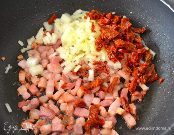 Как только бекон слегка обжариться, добавить к нему нарезанные лук и вяленый помидоры. Перемешать и продолжить обжаривать все вместе еще пару-тройку минут.
