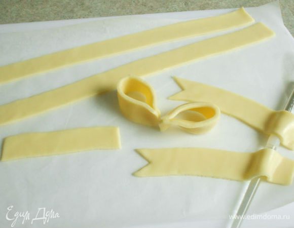 Для банта вырезать 1 полоску 10 х 2,5 см, 2 полоски 11 х 2,5  см и 2 полоски 14 х 2,5  см, вырезая вместе с бумагой и оставляя на ней. У 11 см полосок с одного конца вырезать ножницами треугольники, а противоположные концы уложить на толстую круглую кисточку или фломастер, чтобы сформировать изгиб.Из 14 см полосок сформировать петли банта, прижав концы полосок друг к другу. Снять бумагу и сопоставить концы петель, прижать.
Снять бумагу с 10 см полоски и обернуть ею место склеивания петель. Поставить петли банта вертикально, расправить и убрать в холодильник.
