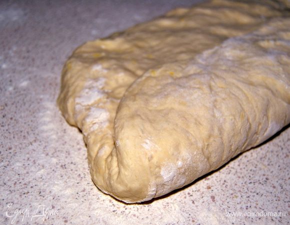 Поднявшиеся тесто выложить на присыпанную мукой поверхность.Расправить в виде прямоугольника.Сформировать колбаску.Для этого завернуть одну из сторон разглаженного теста к середине.Прижмите и запечатайте сгиб.