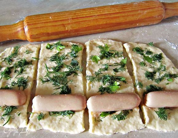 Присыпать слегка мукой стол. Раскатать тесто толщиной около 0,5 мм, нарезать полоски по длине сосисок.  Каждую полоску теста посыпаем зеленью. На край укладываем сосиску и сворачиваем, формируя пирожок.