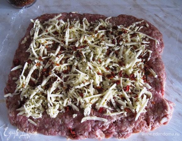 Расстелите на столе кусок пищевой пленки и распределите на нем равномерно фарш. Сверху посыпьте его тертым чеддером со специями и сушеными помидорами.
С помощью пленки аккуратно скрутите фарш в рулет,