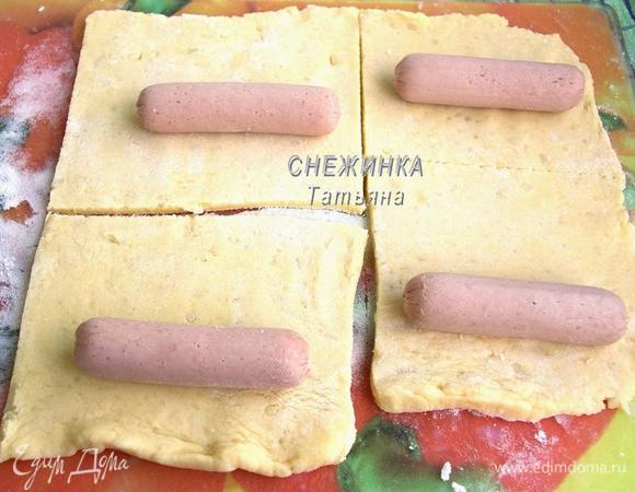 Тесто делим пополам. Раскатываем одну часть в не очень тонкий пласт, толщиной где-то 0,5 мм. Укладываем сосиски, нарезаем тесто на квадраты, чуть больше размера сосисок.