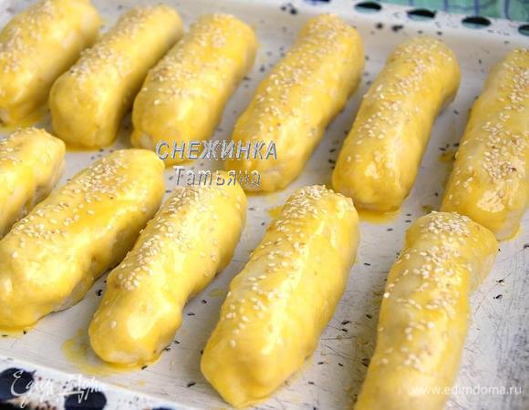 Готовые изделия выкладываем на противень, смазанный растительным маслом. Сливочное масло растапливаем, соединяем с яйцом и хорошо взбиваем вилкой. Смазываем верх и бока изделий полученной смесью. Присыпаем семенем кунжута.