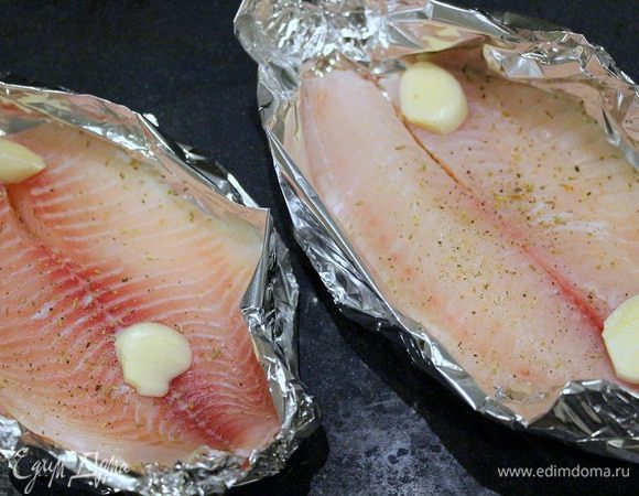 Сделать из фольги лодочки, смазать оливковым маслом тилапию, присыпать травами, посолить, поперчить, уложить в лодочки, сверху положить разрезанные дольки чеснока.