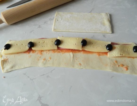 Визуально разделить полоску теста на 2 половинки. Тесто смазать измельчёнными помидорами. На верхнюю половинку теста выложить ветчину, сыр, порезанные оливки, накрыть нижней половиной теста. Так «нафаршировать» все пласты теста.