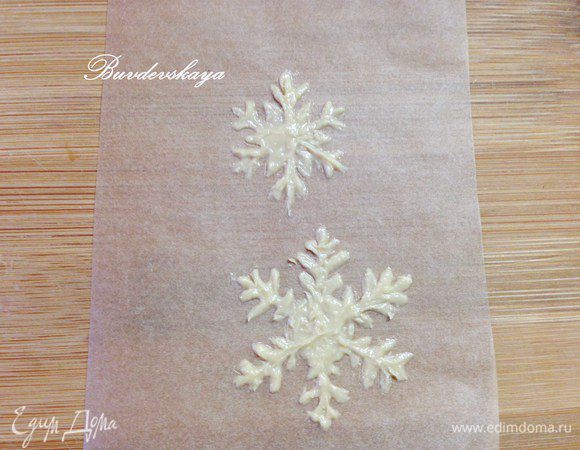 На водяной бане растапливаем белый шоколад, перекладываем в кондитерскую ручку (шприц) или целлофановый пакетик с отрезанным уголком и рисуем на бумаге для выпечки Снежинки. Удобнее это делать по трафарету – распечатать или нарисовать на бумаге снежинки и подложить под пергамент. Можно приклеить их друг к другу двусторонним скотчем, чтобы картинка не смещалась. Пергамент с нарисованными снежинками отправить в морозильную камеру охлаждаться на 20-30 минут.