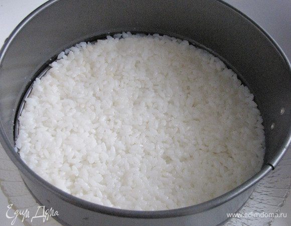 Взять кольцо от разъемной формы для торта  диаметром 18-20 см и поставить на блюдо. Выложить половину риса равномерно, тонким слоем, смачивая руки водой.