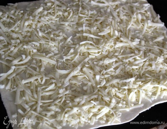 Сыр натрите. Соедините два квадрата слоёного теста в небольшой нахлест. Раскатайте их вместе в одном направлении, но не нарушая слои. Посыпьте поверхность тертым сыром, оставив его немного для оформления при подаче.