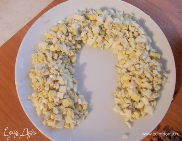 Возьмем плоское блюдо и выкладываем мелко порезанные и сваренные яйца, в форме подковы. Сверху смазываем майонезом.