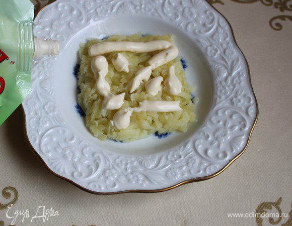 Берем тарелку, я взяла небольшую, поэтому получился маленький салат, но этих продуктов хватит на "Подарок" гораздо большего размера. Картофель натрем на крупной терке. Выкладываем егo на блюдo в форме квадрата. Делаем сверху сеточку из майонезa. Я размазала егo.