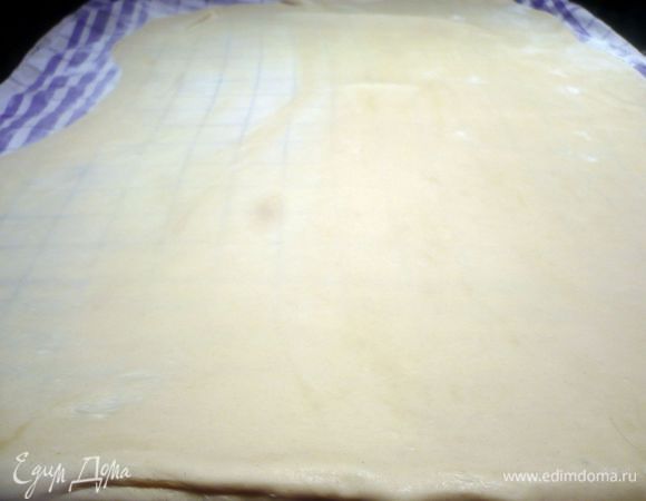 Духовку разогреваем до 200 градусов. Теперь разложим на рабочей поверхности полотенце (коротким краем к себе), присыпем его мукой, скалку также присыпем мукой, тесто извлечем из пленки, выложим тесто на полотенце, припылим его с двух сторон мукой. Начнем раскатывать. Как только тесто будет раскатано почти на всю поверхность полотенца, потянем его снизу по краям на кулачках. На фото видно, что левую сторону я растянула до очень тонкого состояния, правую еще нет. В этот момент я, к счастью, заметила, что на краю, ближнему ко мне, тесто осталось слишком толстым и, если начну раскатывать его, места на полотенце у меня не хватит, тогда эту небольшую часть я отрезала. В связи с этим, лучше выкладывать тесто не совсем у края, а ближе к середине и раскатывать со всех сторон равномерно. 