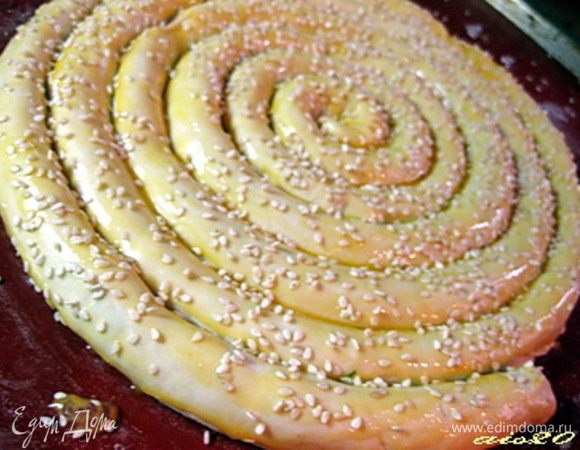 На противень, застеленный бумагой для выпечки, с середины начинаем выкладывать наши жгуты (швом вниз) в виде спирали, скрепляя их между собой. Каждый следующий виток спирали не прислонять слишком близко к предыдущему, чтобы нашему слоеному тесту было место для расслаивания.
Пирог обмазать взбитым яйцом, посыпать кунжутом, сделать несколько проколов вилкой по поверхности пирога, чтобы тесто во время выпечки не вздулось.

