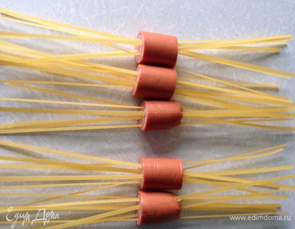 Очистите сосиски от целлофана. Предпочтительнее изделия качественного производства, чтобы не разварились. Разрежьте сосиски на 3-4 части. 
Для одной части понадобится 6-7 макаронин. Просуньте макароны через сосиски так, чтобы они не пересекались, а кусочки расположились посередине. 