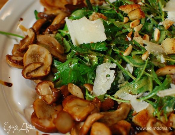 Выложить на блюдо часть листьев руколы, посыпать орехами и сухариками, затем выложить оставшуюся руколу, снова посыпать орехами и сухарями. По краям салата разложить горячие обжаренные грибы, сбрызнуть бальзамическим уксусом, посыпать хлопьями пармезана.