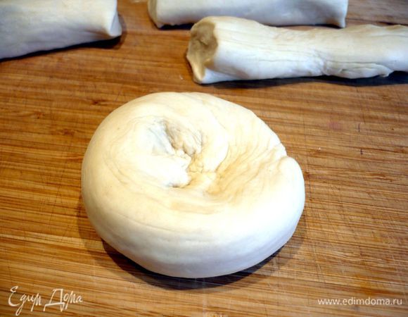 Достаем тесто. Растягиваем в стороны рулет и нарезаем его на 6 частей длиной примерно по 20 см. Каждую часть сворачиваем улиткой и делаем в центре углубление. Раскатываем на присыпанной мукой поверхности в лепешку диаметром примерно 20 см.