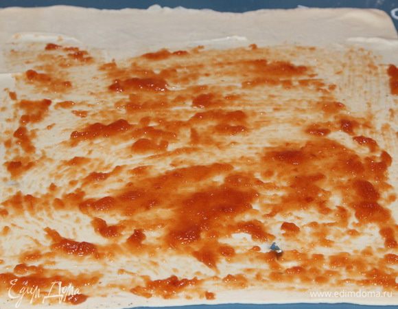 Тесто разделила на две части, расстелила льняное полотенце, подпылила его мукой, выложила тесто и немного раскатала его скалкой. Затем взяла тесто руками и начала его вытягивать в стороны. Тесто очень податливое и вытягивается очень легко. Вытягивайте как можно тоньше, у меня немножко тесто порвалось, но не беда. Разложила тесто на силиконовом коврике и при помощи силиконовой кисточки распределила по поверхности пласта 1 столовую ложку яблочного повидла: 