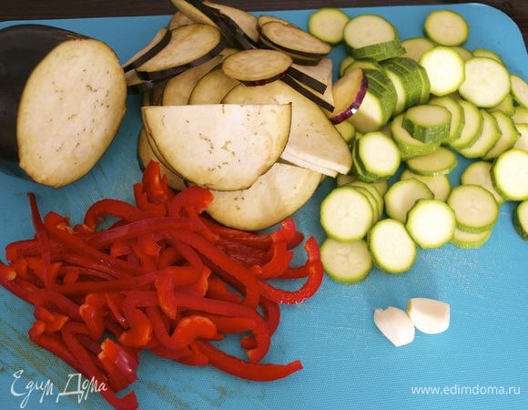 Духовку разогреть до 200 С. Огнеупорную форму для выпечки смазать одной столовой ложкой масла. Все овощи вымыть и подготовить... Обрезать "хвостики"/"ножки"... Кабачок и баклажан нарезать тонкими кружочками. Красный сладкий перец очистить от семян и нарезать тонкими полосками. Чеснок очистить и нарезать пластинами.