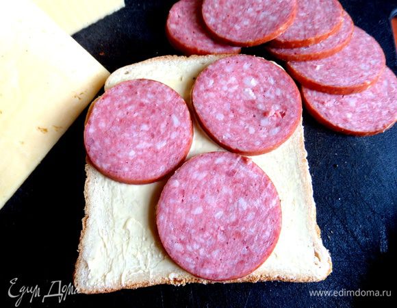 Промазываем маслом хлеб и кладём сверху колбаску. Накрываем квадратиком из сыра.