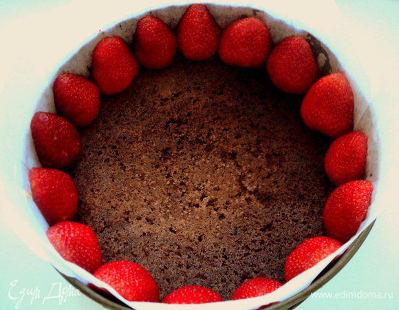 Бока формы выложить бумагой для выпечки, на дно положить шоколадный бисквит. Часть клубники разрезать пополам и установить их  плотно по периметру формы срезом к бокам формы.