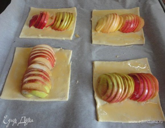 Слоеное тесто (предварительно размороженное) слегка раскатать, чтобы получилось 8 прямоугольников размером на 6 см больше диаметра яблока. У меня были некрупные яблочки. Противень застелить бумагой и выложить тесто. Посередине каждого кусочка выложить «дорожку» из яблок (по 2 половинки одного яблока), края смазать смесью желтка и молока.