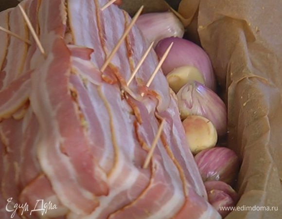 Небольшой глубокий противень выстелить бумагой для выпечки, уложить в него мясо, по бокам разложить луковицы и чеснок.