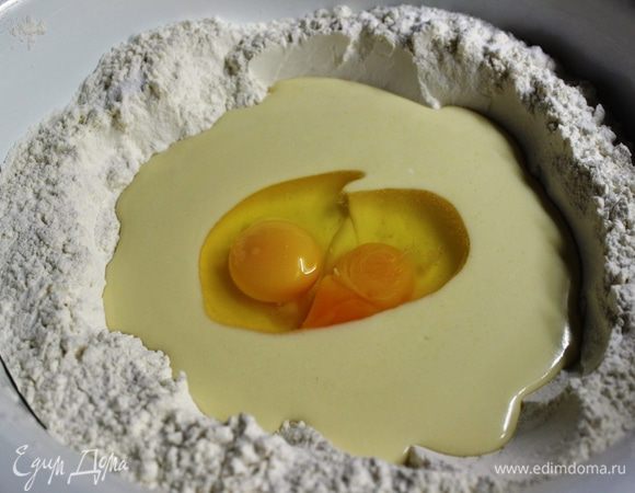 В большое блюдо просеять муку (3/4 части) и добавить разрыхлитель. В центре сделать углубление, вылить в него масляную смесь и разбить два яйца.