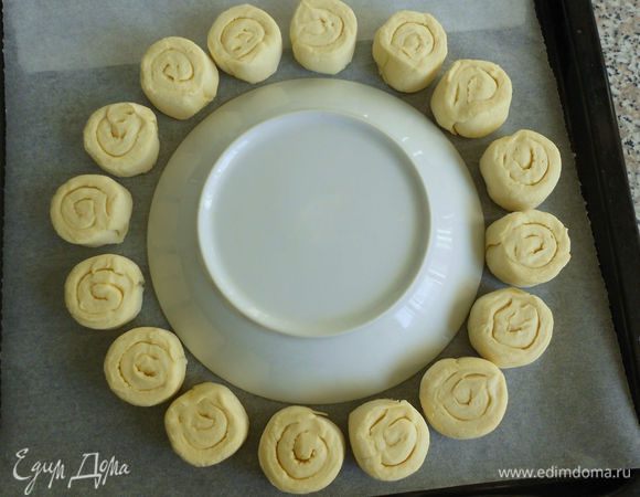 Просторный противень застелите пекарской бумагой. В центр поместите тарелку или крышку от кастрюли диаметром 20 см и выкладываете вокруг 16 заготовок из теста.Тарелка в центре поможет вам создать правильную форму венка. Оставляйте свободным расстояние между булочками и выкладывайте их внешним завитком теста друг к другу, иначе есть риск, что во время выпечки тесто развернётся и форма будет нарушена. Смажьте боковые поверхности заготовок взбитым яйцом.Удалите тарелку с противня и выпекайте булочки в разогретой до 190 градусов С духовке до золотистого цвета.