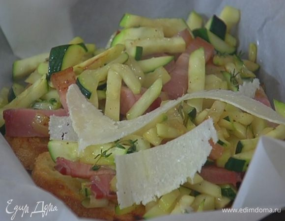 Небольшой противень выстелить бумагой для выпечки, выложить обжаренный хлеб, на него поместить цукини с грудинкой и посыпать хлопьями сыра.