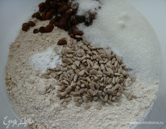 Муку просеять с разрыхлителем. Изюм промыть кипятком, добавить в муку. Всыпать семена подсолнечника и овсяные хлопья. 