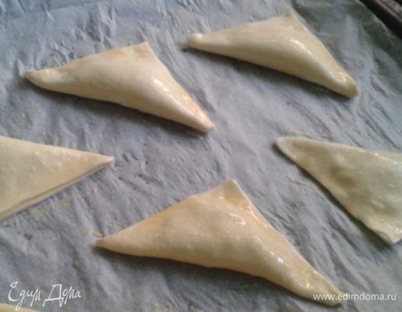 Сложить по диагонали в треугольник и слегка придавить по краю. Заготовки поместить на 15 минут в холодильник. Охлажденные треугольники смазать яичной болтушкой и отправить в горячую духовку. 