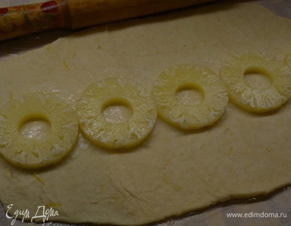 Стол застелить пищевой пленкой (так с тестом лучше работать). Достать тесто и раскатать его в прямоугольный пласт. На середину уложить кружочки ананасов.