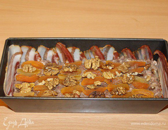 Бекон выложить на дно, курагу разрезать на 2–3 части. Орехи поломать на небольшие кусочки (или вообще не ломать). В форму положить половину фарша. Сверху — орехи и сухофрукты.