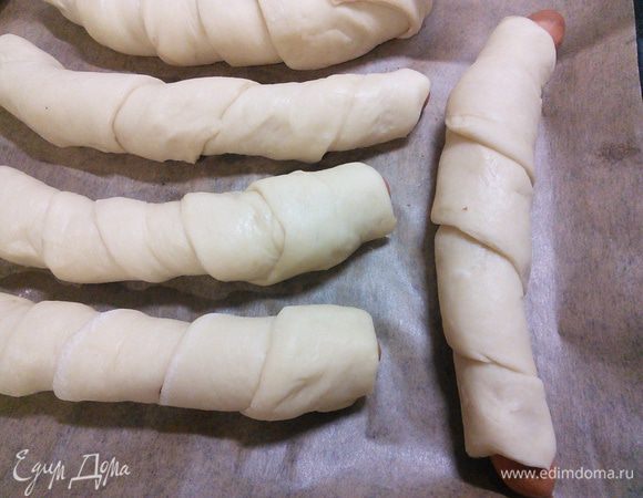 Закрутите каждую сосиску спиралькой в полоски теста, выложите на противень и поставьте на 30 минут в тёплое место на отдых.