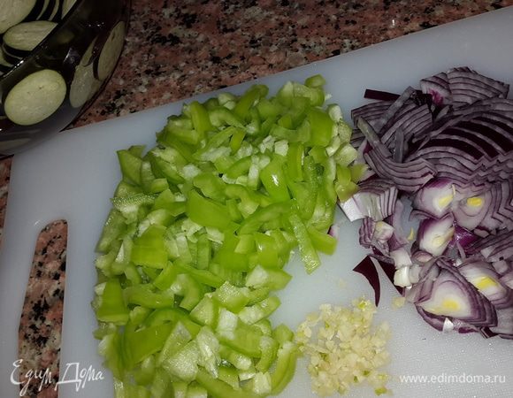 Баклажаны моем, режем на кружочки толщиной не более 0,5 см и замачиваем в холодной воде с солью, чтобы ушла горечь, примерно на 20 минут. Затем промываем под проточной водой и даем обсохнуть. Лук и перец режем на четверть кольца, чеснок мелко рубим. Отправляем лук с чесноком в сковородку с оливковым маслом и обжариваем до мягкости.