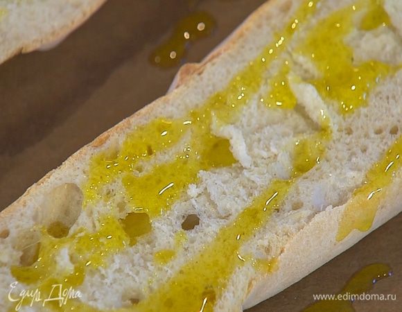 Противень выстелить бумагой для выпечки, выложить багет, сбрызнуть половиной оливкового масла Extra Virgin и смазать томатным соусом.