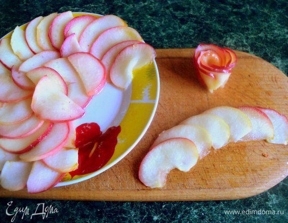 Для украшения яблоки лучше брать краснобокие, тогда розочки получатся яркие и красивые. Яблоки разрезать пополам, удалить семена и нарезать пластинками толщиной 2 мм. Из сахара, воды и лимонного сока сварить сироп. Порциями проваривать в сиропе пластинки яблок буквально несколько секунд, пока они станут мягкими, но не переварятся. Выложить пластинки яблок на тарелку, чтобы стек сироп. Затем свернуть из яблок розочки.