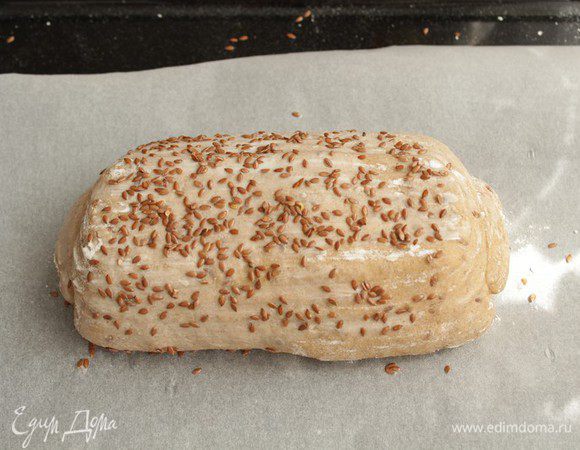 Накройте хлеб, чтобы уберечь от заветривания, и отправьте в теплое место на 50-60 минут. За это время буханка вырастет. Переверните хлеб на противень аккуратно, чтобы воздух не вышел.

Сбрызните буханку водой и посыпьте льном.

