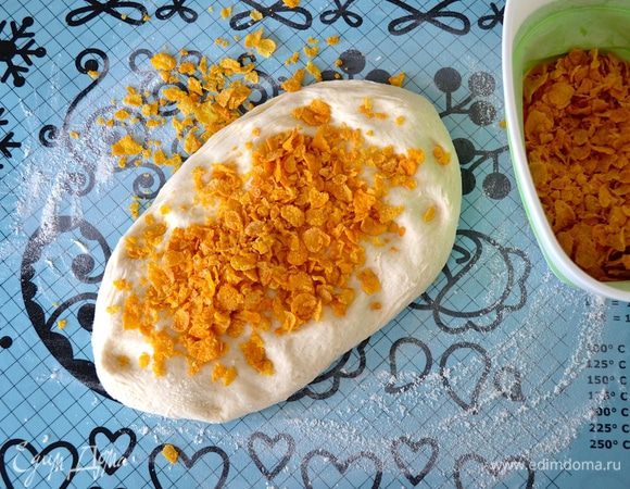 Выложите подошедшее тесто на рабочую поверхность, расплющите слегка и распределите 50-60 г кукурузных хлопьев (несладких). Вымесите еще немного тесто, позволяя хлопьям распределиться по всему тесту. 