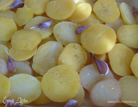 Форму для запекания смазать оливковым маслом, выложить нарезанный кружочками картофель, каждый слой солим и перчим.  Чеснок помыть и не снимая шкурки разбросать поверх картофеля.