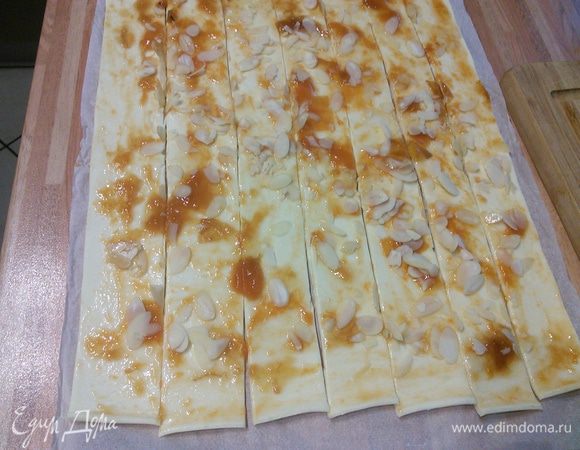 Пласт теста смазать абрикосовым джемом (вареньем, мармеладом), посыпать миндальными лепестками. Разрезать вдоль на полоски шириной около 3 см. 