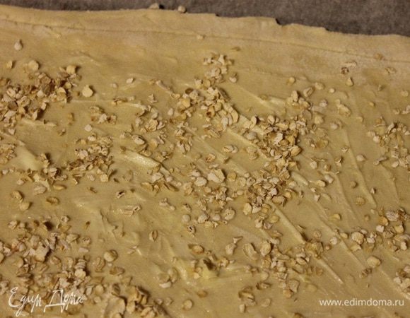 Тесто поделить на две неравные части. Большую часть равномерно выложить в форму для выпечки, смазать сливочным маслом и посыпать овсяными хлопьями.