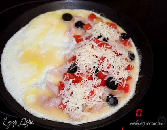 Как только края у омлета схватятся, а середина омлета еще слегка жидкая, на одну половину омлета выложить всю начинку — колбасу, томат, оливки и сыр.