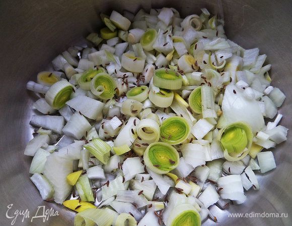 Берём кастрюлю с толстым дном, наливаем масло, высыпаем кольца порея, измельчённый репчатый, тмин, ставим на огонь. Немного обжарим.