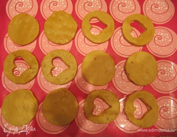 На слегка присыпанной мукой поверхности раскатайте тесто. С помощью круглой формочки (диаметром 5–6 см) вырежьте из теста кружки. В половине кружков с помощью формочки маленького размера любой формы (у меня — сердечко) вырежьте отверстия по центру для того, чтобы в готовом печенье была видна начинка. Выложите заготовки для печений на противень, застеленный силиконовым ковриком или бумагой для выпечки.<br />
Тесто достаточно хрупкое, поэтому удобнее раскатывать в пласт не все тесто сразу, а отщипывать небольшой кусочек, раскатывать его и затем вырезать кружок с помощью формочки. Центральные отверстия лучше вырезать, когда цельные кружки уже выложены на противень.