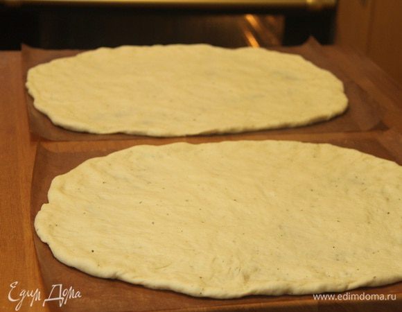 Тесто выложить на рабочую поверхность, разделить пополам. Из каждого куска сформировать основу пиццы (я делаю это руками, диаметр 30 см). Выложить основы на антипригарные листы. Оставить в покое минут на 15.
Прогреть духовку до максимума (у меня 250°С) вместе с противнями.