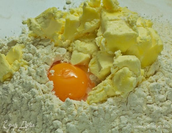 Муку просеять горкой, в углубление всыпать пудру, добавить яичный желток, нарубленное мелкими кубиками масло. Размять руками, начать замешивать, примешивая муку от краев к центру.